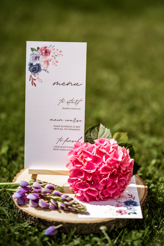Menu card with floral design on a wooden stand with pink flowers and greenery in the grass